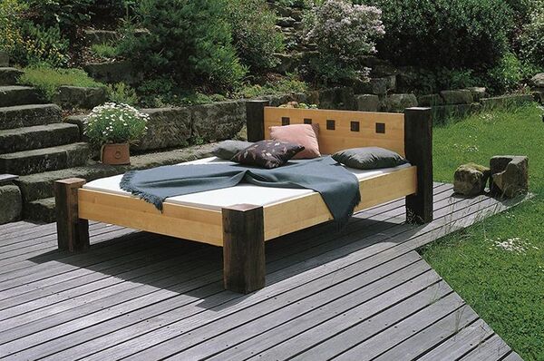 Modernes Holzbett mit Kissen und Decke auf Terrasse im Garten mit Natursteinmauer.